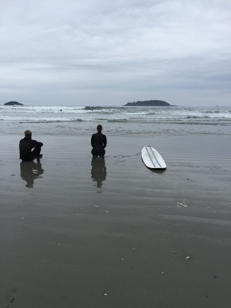 two people sitting on a sand beach with waves coming in and a surfboard on the sand next to them.